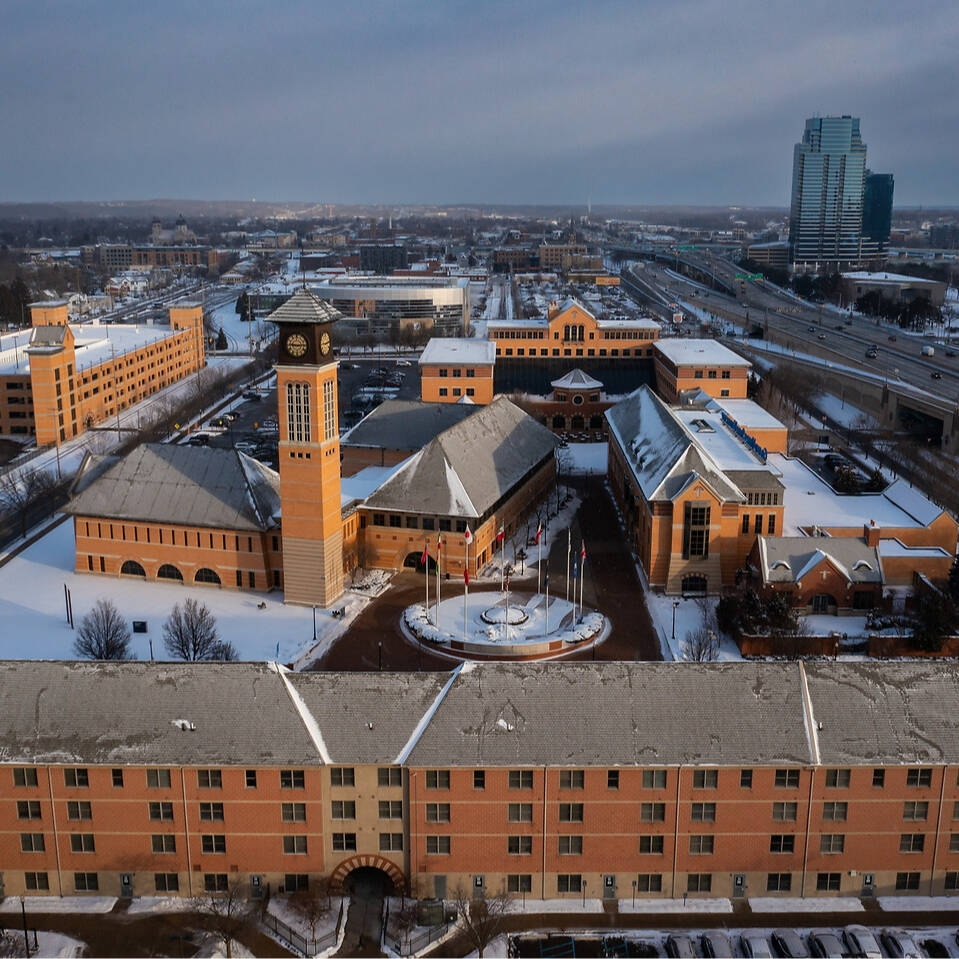 An aerial view of a snow-covered urban complex featuring a prominent clock tower and surrounding buildings, with a highway and modern skyscraper visible in the distance.